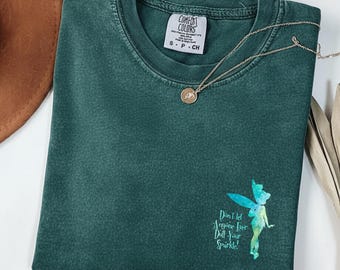 May include: A dark green short-sleeve t-shirt with a crew neckline and a Comfort Colors brand label. The shirt features a watercolor-style graphic of a fairy in shades of blue and green, accompanied by the text, Don't let anyone ever dull your sparkle!