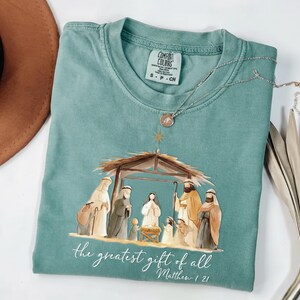 May include: A teal Comfort Colors t-shirt featuring a Nativity scene illustration. The graphic depicts figures in a stable, with the text "the greatest gift of all" and Matthew 1:21. A silver necklace with a pendant rests on the shirt.