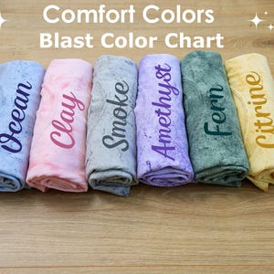 A collection of six rolled-up towels or shirts, each with a different color printed on them, arranged on a wooden surface.