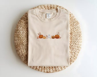 May include: A beige crew neck t-shirt featuring a central embroidered graphic of three orange pumpkins, a small yellow pumpkin, and a white ghost, all surrounded by brown autumn leaves and small berries, arranged in a horizontal line across the chest.