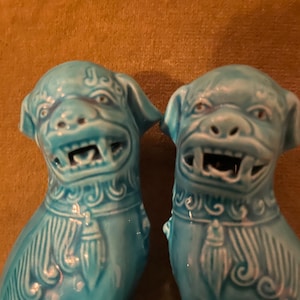 May include: Two turquoise ceramic Foo Dog statues. Each dog has a detailed mane, collar, and a fierce expression with visible teeth. The statues are set against a brown background.