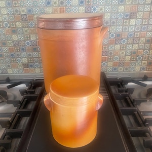 May include: Two brown ceramic jars with lids, the smaller jar is sitting on top of the larger jar. The jars are on a black surface.