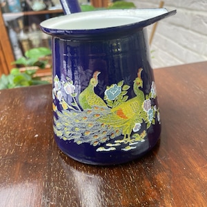 May include: A blue enamel jug with a white interior and a handle. The jug features a painted design of two peacocks with yellow, green, and white feathers.