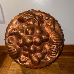 May include: A round copper mould featuring a detailed embossed design of a cluster of grapes with leaves. The metallic surface has a warm, reddish-brown tone with a hammered texture and visible indentations that define the fruit and foliage.