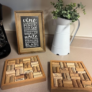 May include: Two wooden coasters with a square shape and a black frame. Each coaster is filled with a grid of used wine corks. The corks have various labels and logos, including "19 Crimes", "DAOU", "BERINGER", "FRANCISCAN", "MAGISTRATE", "MILESTONE", "COPPOLA", and "THIE". Above the coasters is a framed black sign with white text that reads "vino CABERNET PINOT NOIR red ZINFANDEL SANGIOVESE MERLOT white PINOT GRIGIO RIESLING SAUVIGNON BLANC CHARDONNAY".