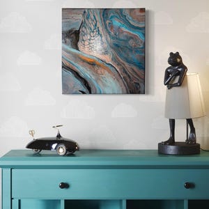 May include: A teal blue dresser with three drawers and two shelves. A black toy car sits on the top of the dresser. A frog lamp with a gray lampshade sits on the right side of the dresser. A framed abstract painting with blue, brown, and white colors hangs on the wall behind the dresser.