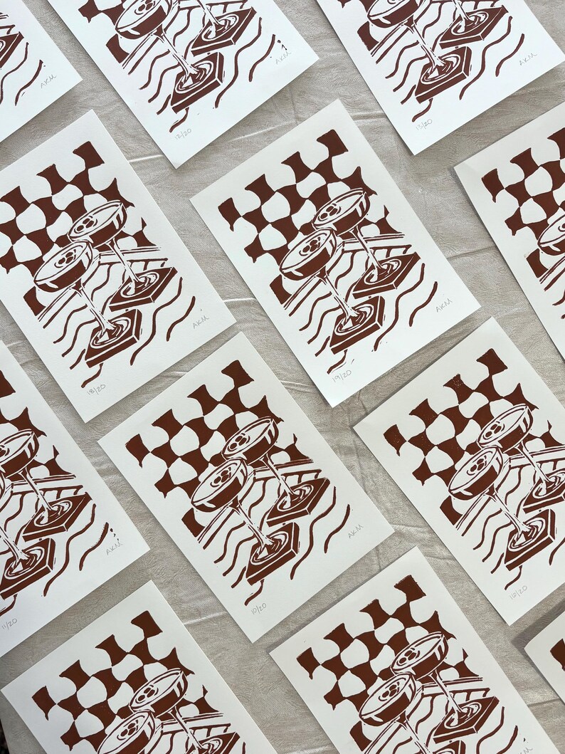 Puede incluir: Una colecci&oacute;n de grabados art&iacute;sticos en blanco y marr&oacute;n. Cada grabado presenta una ilustraci&oacute;n estilizada de copas de c&oacute;ctel, un patr&oacute;n de cuadros y l&iacute;neas onduladas. Los grabados est&aacute;n numerados y firmados, con el texto "AKM" visible.