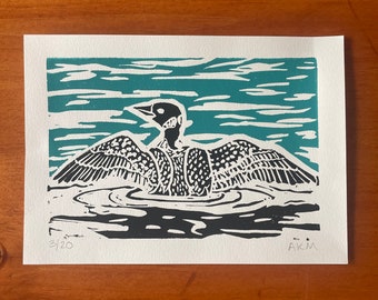 May include: A black and white linocut print of a loon swimming in teal water. The print is numbered 3/20 and signed "AKM".