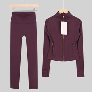 May include: A matching athletic set in a deep burgundy color. The set includes high-waisted leggings and a zip-up jacket with gold-toned zippers. Both items are displayed on wooden hangers against a neutral background.