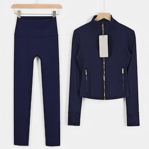 May include: A navy blue athletic set, including high-waisted leggings and a zip-up jacket. The jacket has a stand-up collar, gold zipper, and two front pockets. The set is displayed on wooden hangers against a white background.