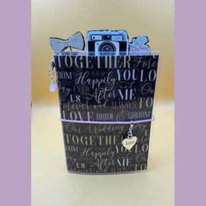 May include: A black notebook cover featuring white wedding-themed text, including the words Together, Happily, and Bride & Groom. It is adorned with a white bow charm, a camera cutout, a floral accent, and a silver heart charm labeled love.