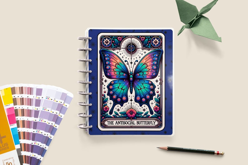 May include: A blue and white spiral bound notebook featuring a colourful butterfly illustration on the cover. The butterfly has iridescent wings with blue, green, and pink hues. The text "THE ANTISOCIAL BUTTERFLY" is printed below the butterfly.