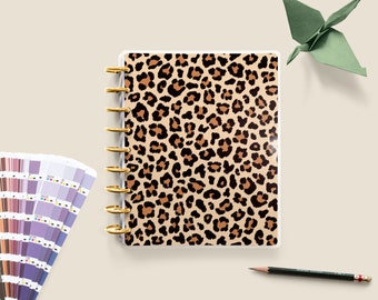 May include: A clear plastic planner with a leopard print design. The planner has gold rings and is lying on a beige surface.