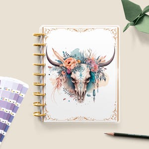 May include: A white planner with a gold spiral binding features a watercolor illustration of a skull with a floral crown. The skull is surrounded by feathers and other decorative elements.