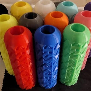 May include: A collection of colorful, 3D-printed bottle stoppers. The stoppers are in various colors, including red, yellow, blue, green, pink, and purple. Each stopper has a unique geometric design and a rounded top.