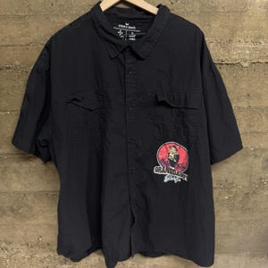 May include: Black short-sleeved button-down shirt with two chest pockets. The shirt features a graphic on the lower right with the text "GRANDDADDY SHOP" and an illustration of a man. The shirt has the "STEVE'S JEANS" label.