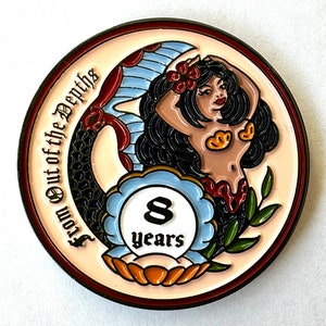 Op de afbeelding: Een ronde, kleurrijke munt met een zeemeermin met zwart haar en een rode bloem. De munt heeft de tekst "From Out of the Depths" en "8 years". De zeemeermin wordt omringd door een blauw en wit ontwerp.