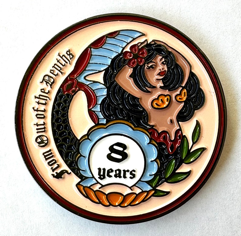 Op de afbeelding: Een ronde, kleurrijke munt met een zeemeermin met zwart haar en een rode bloem. De munt heeft de tekst "From Out of the Depths" en "8 years". De zeemeermin wordt omringd door een blauw en wit ontwerp.