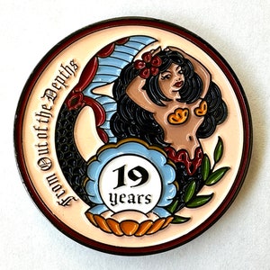 Op de afbeelding: Een ronde, kleurrijke pin met een zeemeerminontwerp. De zeemeermin heeft zwart haar, rode bloemen en een rode en blauwe staart. De pin heeft de tekst "From Out of the Depths" en "19 years".