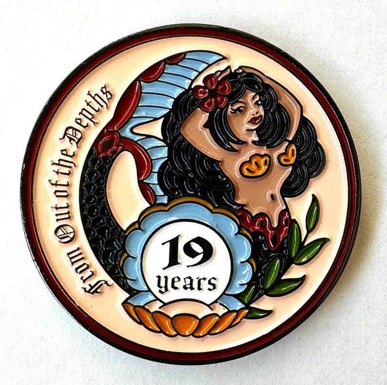 Op de afbeelding: Een ronde, kleurrijke pin met een zeemeerminontwerp. De zeemeermin heeft zwart haar, rode bloemen en een rode en blauwe staart. De pin heeft de tekst "From Out of the Depths" en "19 years".