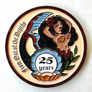 Op de afbeelding: Een ronde, decoratieve pin met een zeemeerminontwerp. De zeemeermin heeft zwart haar, een gebruinde teint en een rood en goud schelpen topje. De pin heeft de tekst "From Out of the Depths" en "25 years".