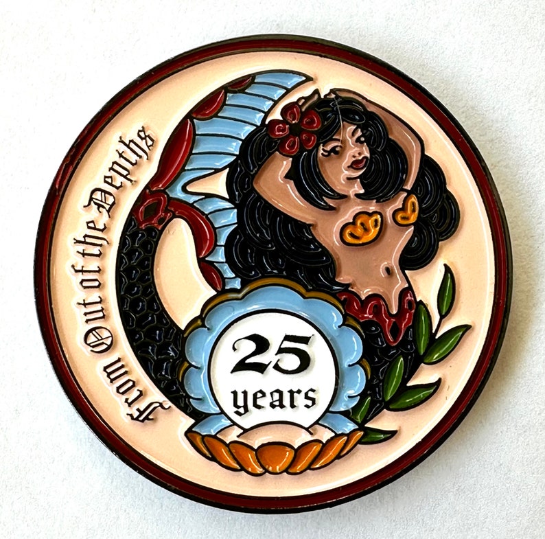 Op de afbeelding: Een ronde, decoratieve pin met een zeemeerminontwerp. De zeemeermin heeft zwart haar, een gebruinde teint en een rood en goud schelpen topje. De pin heeft de tekst "From Out of the Depths" en "25 years".