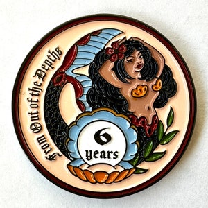 Op de afbeelding: Een ronde, decoratieve munt met een zeemeermin met zwart haar en een rode bloem. De munt heeft de tekst "From Out of the Depths" en "6 years". De zeemeermin heeft een bruine teint en is omgeven door een blauw en rood ontwerp.