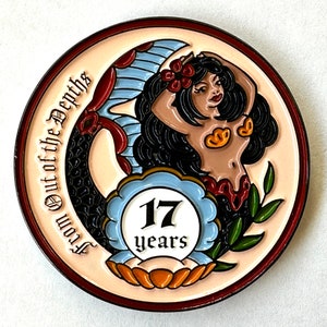 Op de afbeelding: Een ronde, cr&egrave;mekleurige pin met een zeemeerminontwerp. De zeemeermin heeft zwart haar, een gouden top en een rode rok. De pin heeft de tekst "From Out of the Depths" en "17 years" in een blauw-wit embleem.
