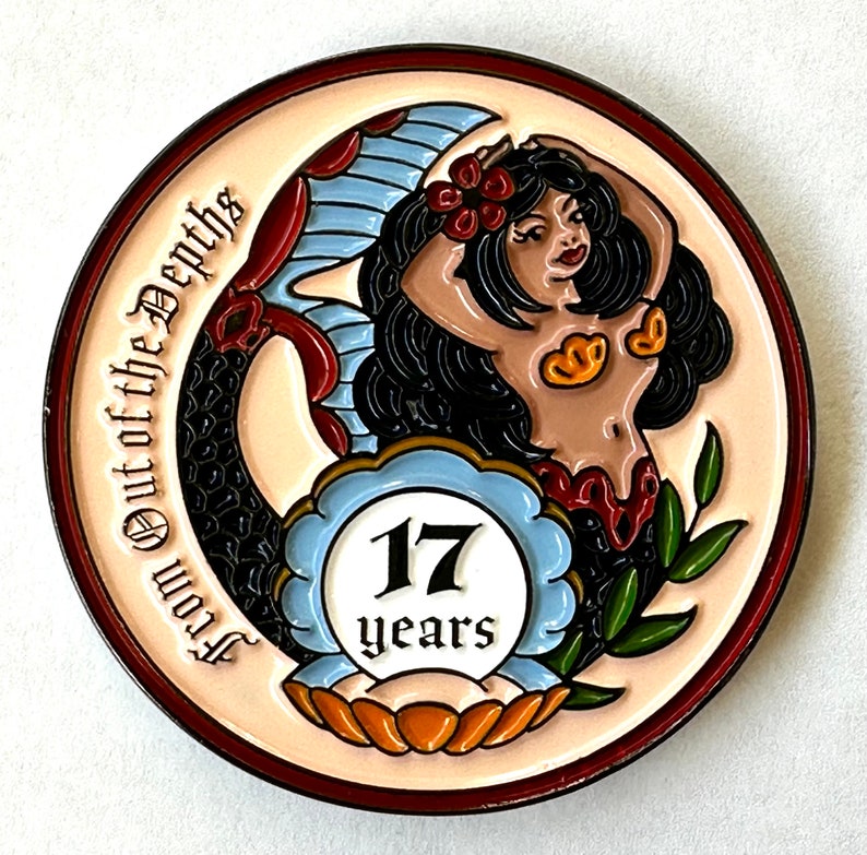 Op de afbeelding: Een ronde, cr&egrave;mekleurige pin met een zeemeerminontwerp. De zeemeermin heeft zwart haar, een gouden top en een rode rok. De pin heeft de tekst "From Out of the Depths" en "17 years" in een blauw-wit embleem.