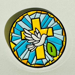 May include: A round, colorful emblem featuring a yellow cross, a white dove, and a green leaf with the number 1. The design is reminiscent of stained glass, with blue, white, and yellow geometric shapes.