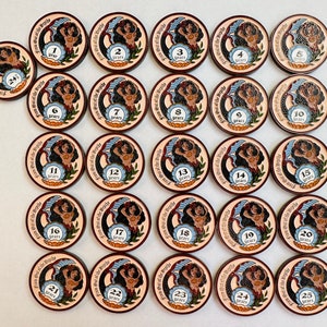 Op de afbeelding: Een verzameling ronde, houten tokens met een zeemeermin illustratie. Elke token heeft een nummer van 1 tot 25, met de tekst "From Out of the Depths" en "years" langs de rand. De tokens zijn bruin, beige en blauw.