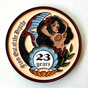 Op de afbeelding: Een ronde, cr&egrave;mekleurige pin met een zeemeerminontwerp. De zeemeermin heeft zwart haar, een rode bloem en gouden schelpaccenten. De pin heeft de tekst "From Out of the Depths" en "23 years" in een blauw-wit embleem.