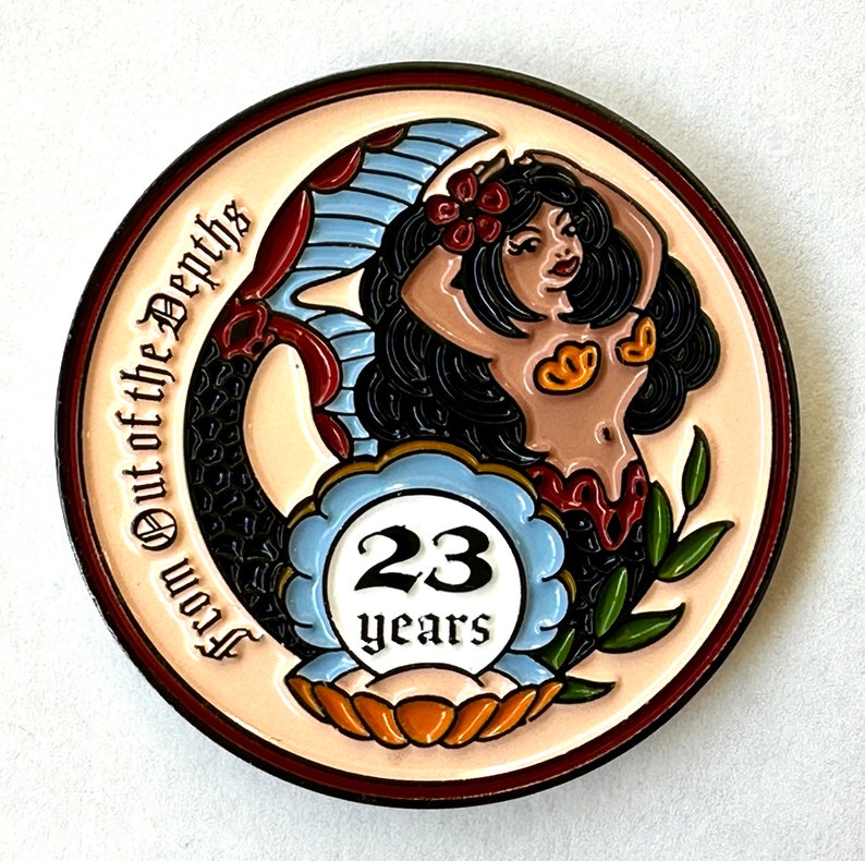Op de afbeelding: Een ronde, cr&egrave;mekleurige pin met een zeemeerminontwerp. De zeemeermin heeft zwart haar, een rode bloem en gouden schelpaccenten. De pin heeft de tekst "From Out of the Depths" en "23 years" in een blauw-wit embleem.