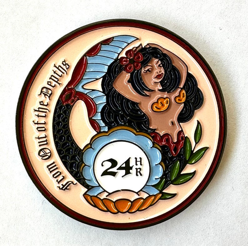 Op de afbeelding: Een ronde, kleurrijke munt met een zeemeerminontwerp. De zeemeermin heeft zwart haar, rode bloemen en een schelpvormige top. De munt heeft de tekst "From Out of the Depths" en "24HR".