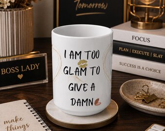 May include: A white ceramic mug featuring the black text I AM TOO GLAM TO GIVE A DAMN, accented with a gold-colored lip print and a small pink heart icon. The mug sits on a circular marble-patterned coaster with a gold-toned metal rim.