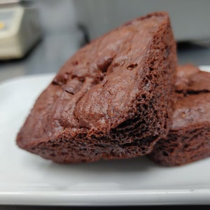 May include: Two dark chocolate brownies on a white plate. The brownies have a rich, porous texture and a deep brown colour. The image is a close-up, highlighting the details of the baked goods.