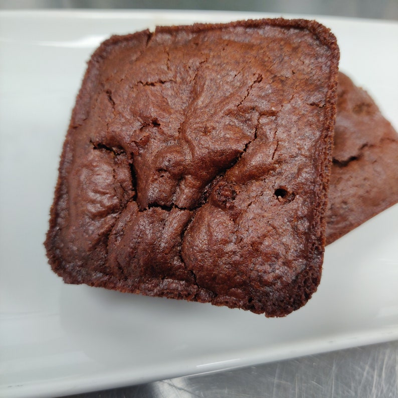May include: Two square chocolate brownies on a white plate. The brownies are a deep brown colour with a slightly cracked surface. The edges are darker, indicating a baked texture. A simple, appetizing presentation of a classic treat.