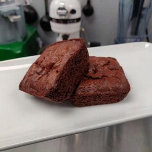 May include: Two square, dark brown brownies on a white rectangular plate. The brownies have a slightly textured surface and appear freshly baked. Kitchen appliances are visible in the background.