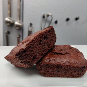 May include: Two dark brown brownies on a white plate. One is cut, showing the porous interior. The other is whole. The background is a light grey with kitchen utensils hanging on the wall.