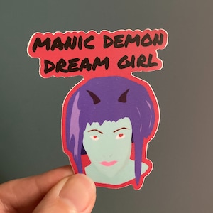 May include: A sticker featuring a cartoon illustration of a woman with purple hair and red horns. The text "MANIC DEMON DREAM GIRL" is written in black on a pink background.