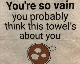 Könnte beinhalten: Weißes Küchentuch mit schwarzem Text, der "You're so vain you probably think this towel's about you" lautet. Unter dem Text ist eine braune Kaffeetasse mit weißen Wolken gezeichnet.