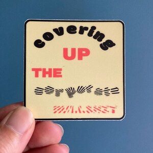 May include: A yellow sticker with black and red text that reads "covering up the corporate planet".