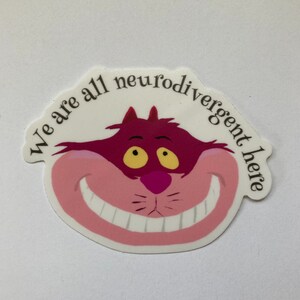 May include: A white sticker featuring a pink Cheshire Cat from Alice in Wonderland. The cat has a wide grin and yellow eyes. The text surrounding the cat reads "We are all neurodivergent here."
