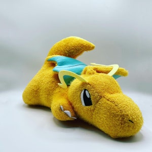 May include: A yellow plush toy dragon with a blue and yellow wing. The dragon has a friendly face with black eyes and a yellow horn.