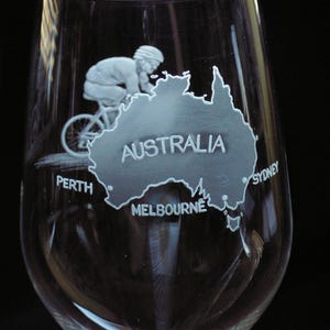Puede incluir: Una copa de vino de vidrio transparente con un diseño grabado de un ciclista que recorre un mapa de Australia. El mapa incluye las ciudades de Perth, Melbourne y Sydney.