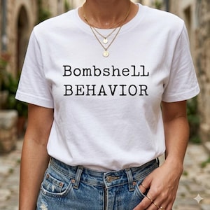 May include: White t-shirt with the text "Bombshell BEHAVIOR" in black. The shirt is paired with blue denim jeans. A gold necklace with layered chains and pendants is worn.