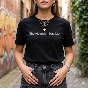May include: A black short-sleeved t-shirt featuring the white text The Algorithm Sent Me printed across the chest. The shirt is paired with dark gray denim jeans, gold layered necklaces, gold hoop earrings, and gold rings on the fingers.