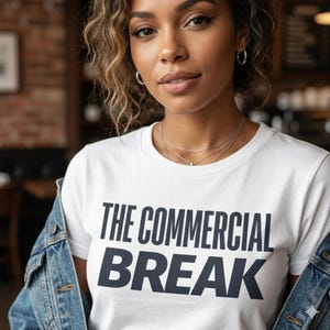 May include: White t-shirt with the text "THE COMMERCIAL BREAK" in dark blue block letters. The person is wearing a denim jacket and silver necklaces. The background is blurred.