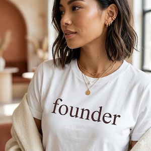 Op de afbeelding: Wit t-shirt met het woord "founder" in donkerbruin gedrukt. De persoon draagt gouden kettingen en oorbellen. Een beige jasje is over de schouders gedrapeerd. De focus ligt op het t-shirt.