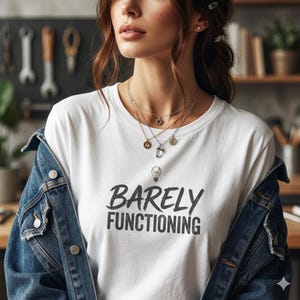 Puede incluir: Camiseta blanca con el texto "Barely Functioning" en gris oscuro. La persona lleva una chaqueta vaquera y collares superpuestos con pequeños colgantes. El fondo incluye herramientas y una estantería de madera.